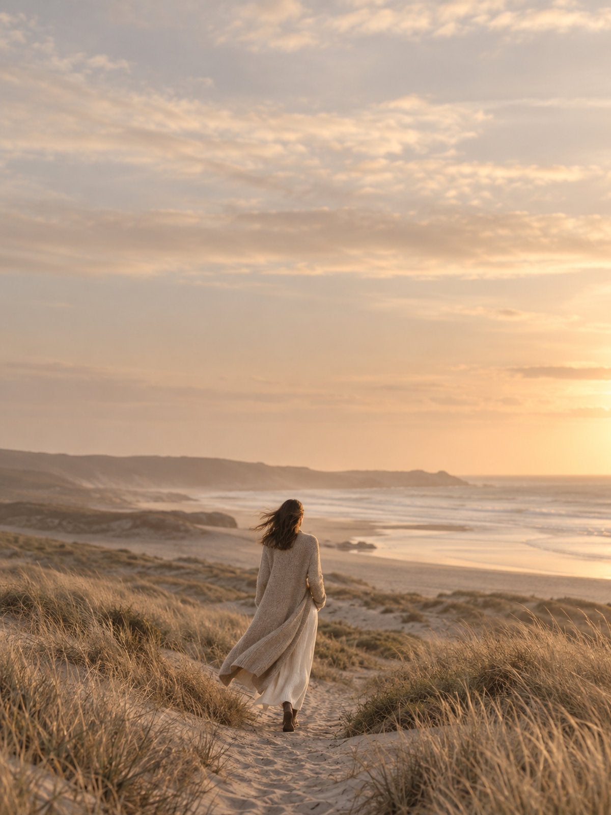 Woman in a long knit cardigan walking along the coast at golden hour